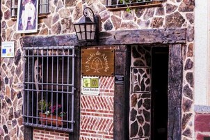A stone building with a rustic entrance, a wooden signboard, and a barred window with potted plants.