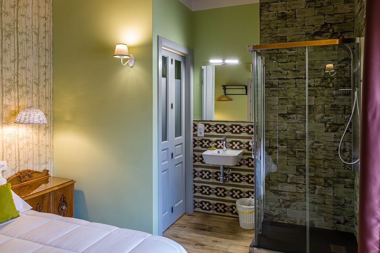 A bedroom with a bed, bedside table, lamp, and a bathroom with a shower, sink, and patterned tiles.