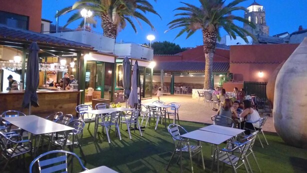 Un espace extérieur aménagé avec des tables et des chaises, des palmiers et un bâtiment abritant un bar.
