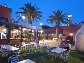 An outdoor seating area with tables and chairs, palm trees, and a building with a bar.