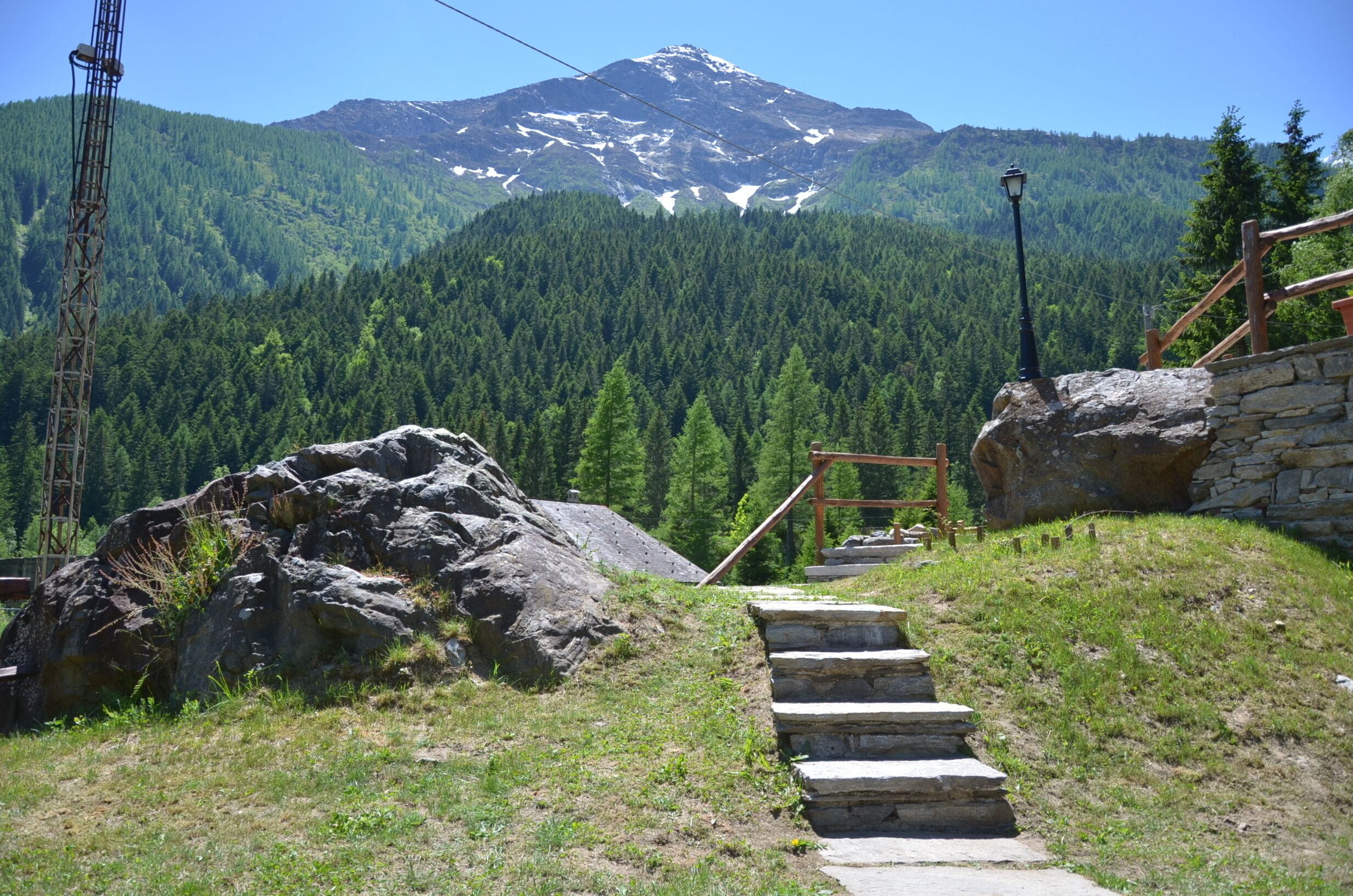 Sendero de montaña con escalones de piedra que ascienden, rodeado de un bosque verde exuberante y un pico nevado al fondo.