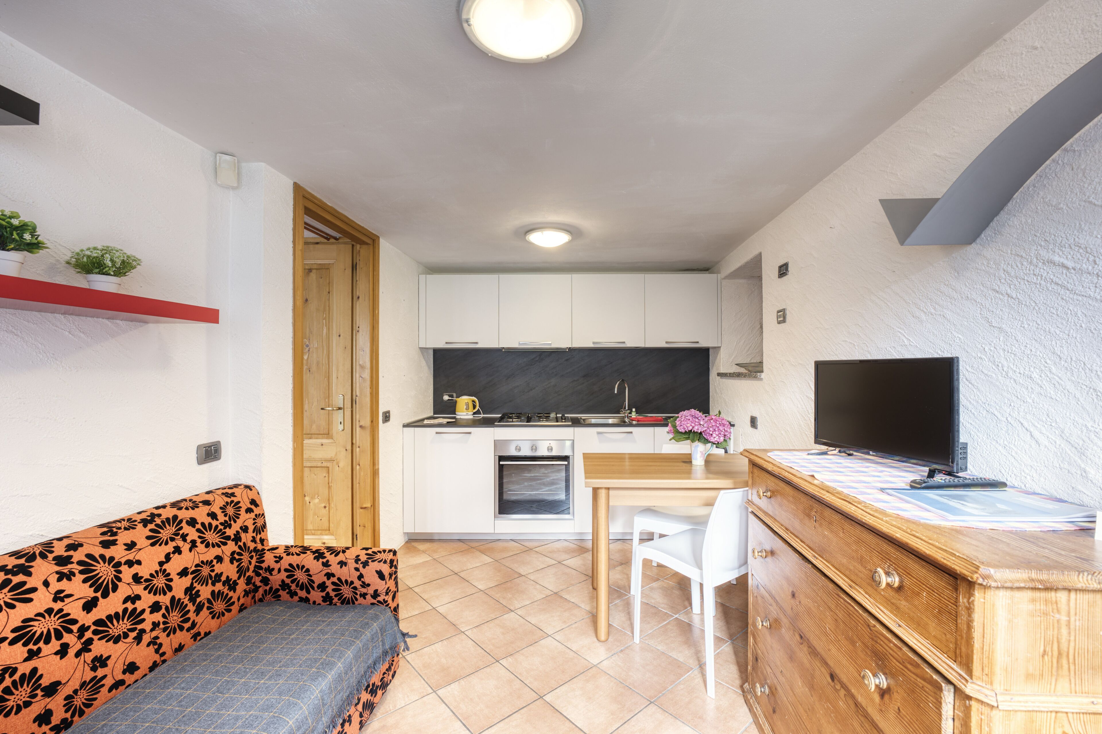 A compact kitchen with white cabinets, a wooden countertop, and a built-in oven. There is a dining table with a chair, a sofa with a floral pattern, and a television on a wooden dresser.