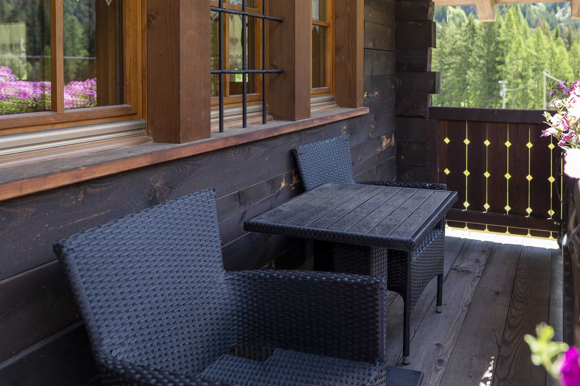 A wooden deck with two wicker chairs and a table, a wooden railing, and a window with a view of trees.