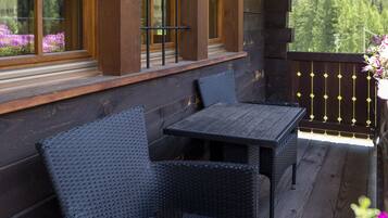 A wooden deck with two wicker chairs and a table, a wooden railing, and a window with a view of trees.