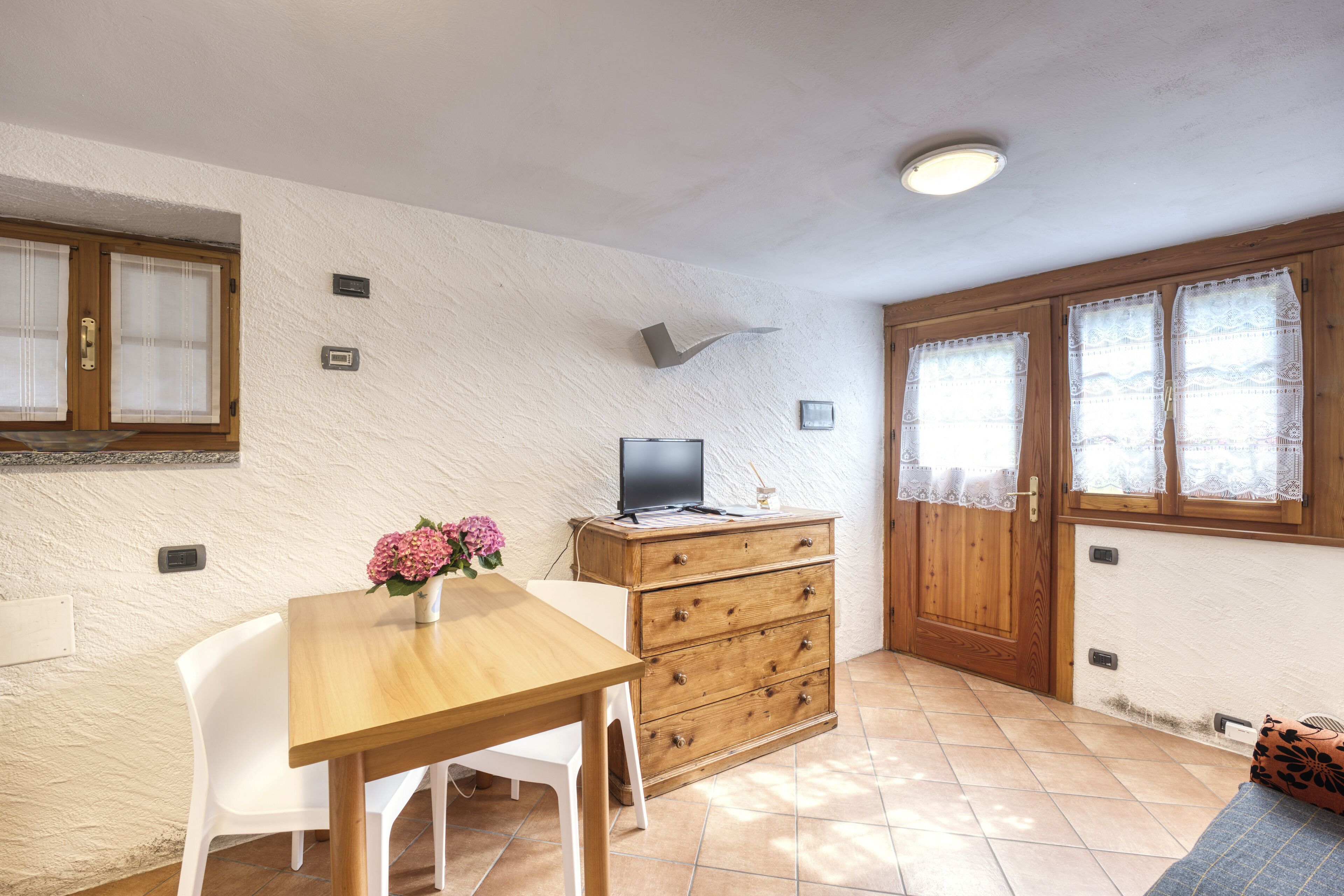 Una habitación pequeña con una mesa de madera, una silla blanca, un armario de madera y un televisor encima.