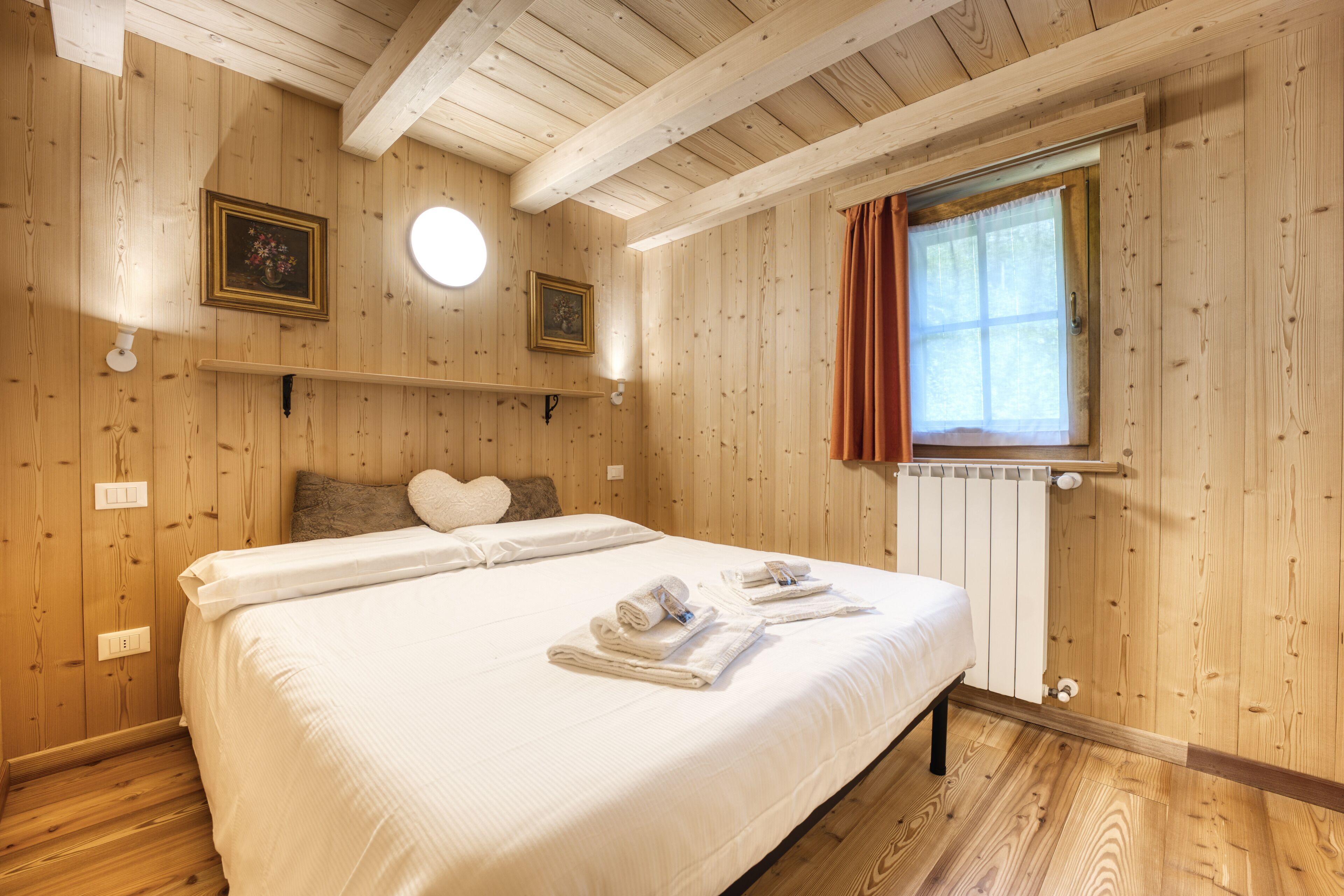 Un dormitorio con paneles de madera, una cama, dos lámparas fijadas a la pared, un ventanal con cortinas y un radiador.