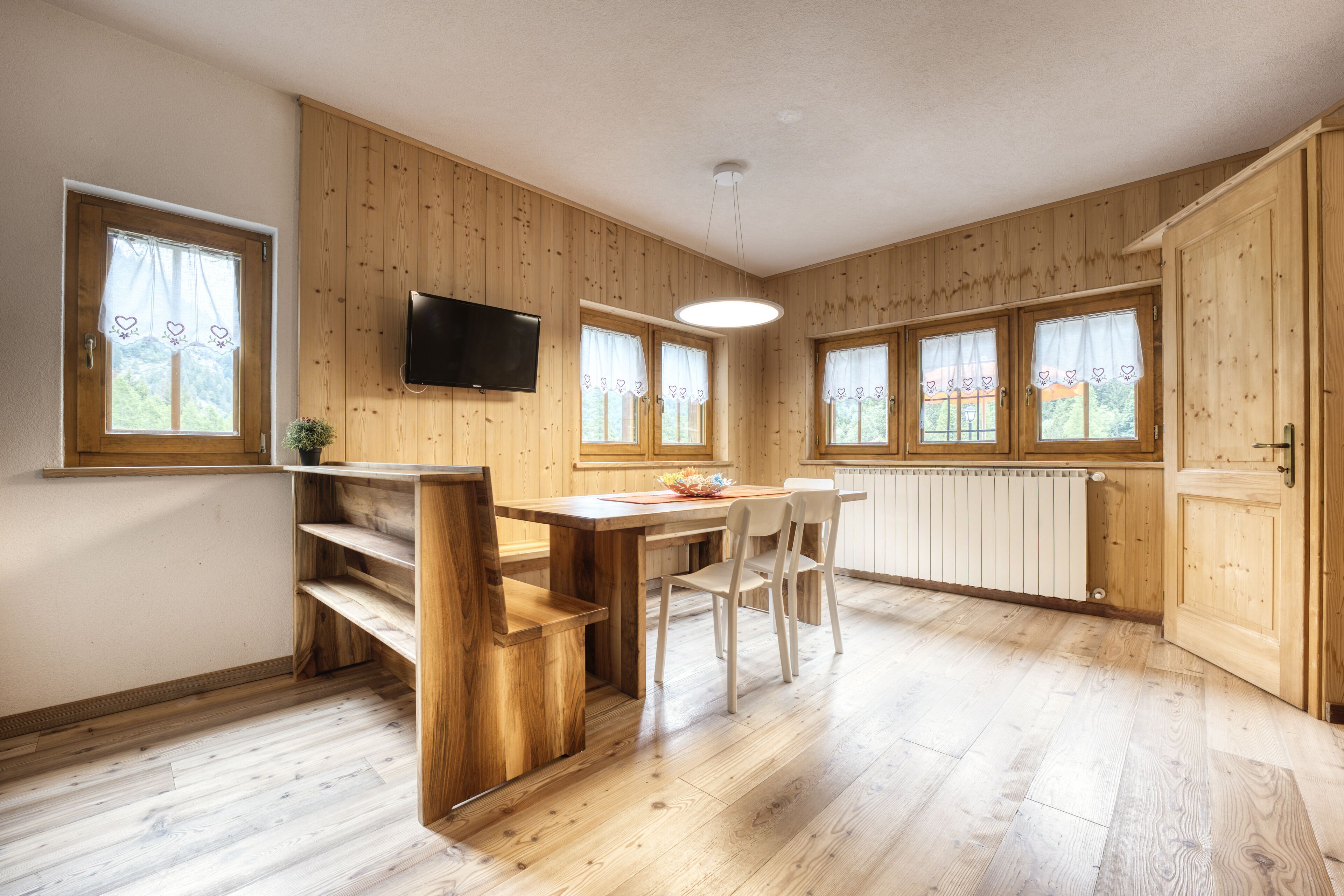 Ein Zimmer mit Holzwandverkleidung, einem Esstisch mit Stühlen, einem an der Wand montierten Fernseher und großen Fenstern mit Vorhängen.