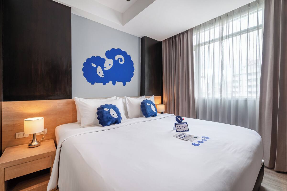 A hotel room with a large bed, white bedding, a wall-mounted sheep decoration, a nightstand with a lamp, and a window with curtains.