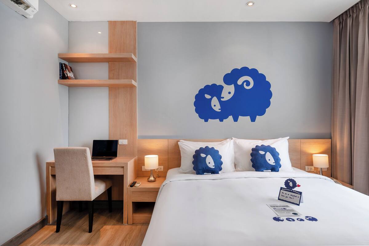A hotel room with a bed, a desk with a laptop, a chair, and a wall with a blue sheep decoration.