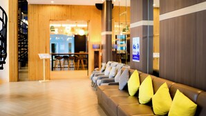 A modern hotel lobby with a wooden reception desk, a seating area with a brown sofa and yellow cushions, and a herringbone-patterned floor. - Kokotel Bangkok Dheva Thonglor (Bangkok)