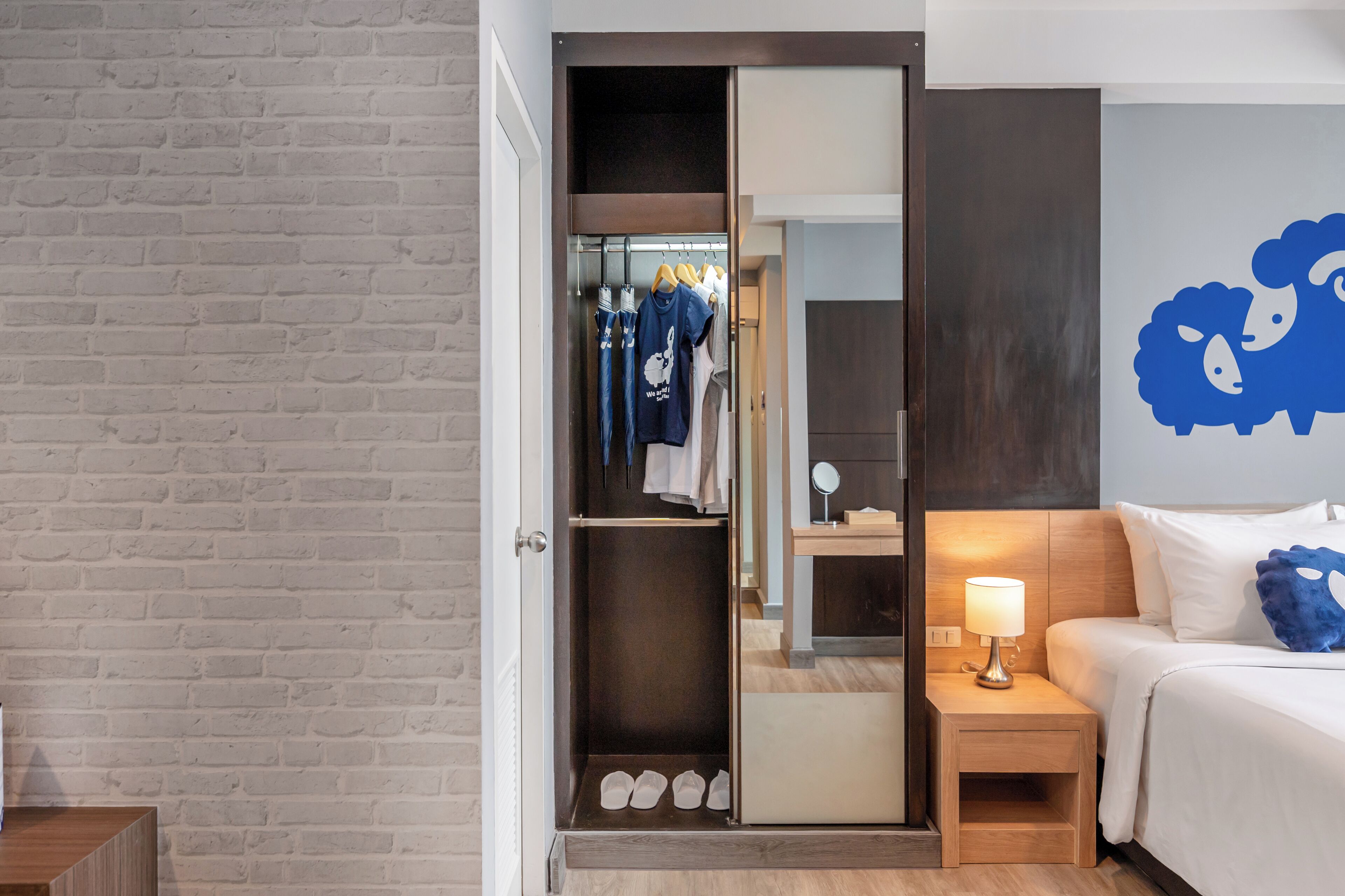 A hotel room with a brick wall, a wardrobe with hanging clothes, a bedside table with a lamp, and a bed with white bedding and a blue pillow.