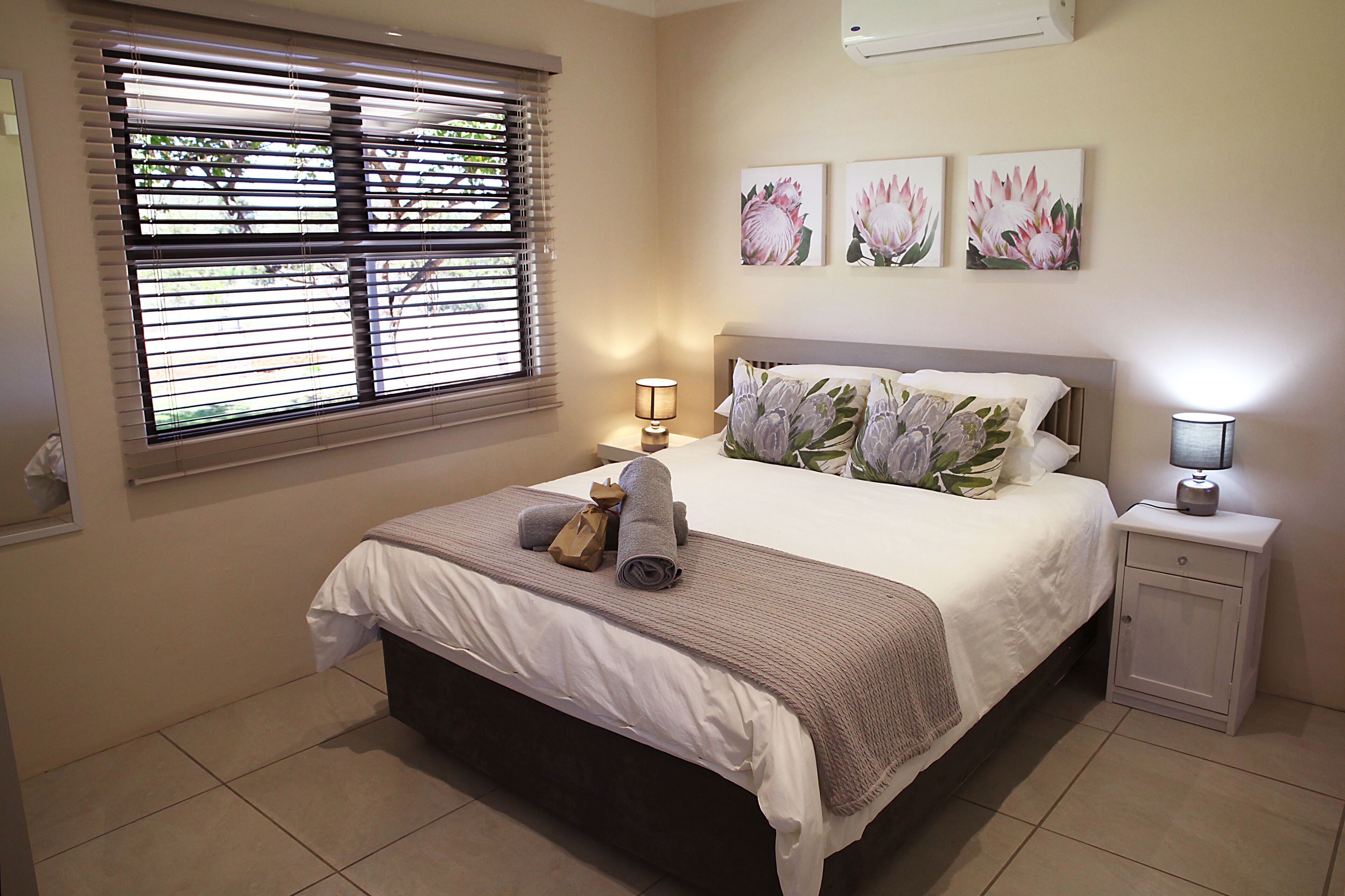 A neatly made bed with a white and gray duvet, a bedside lamp, and a nightstand with a lamp. There are three floral wall art pieces above the bed.