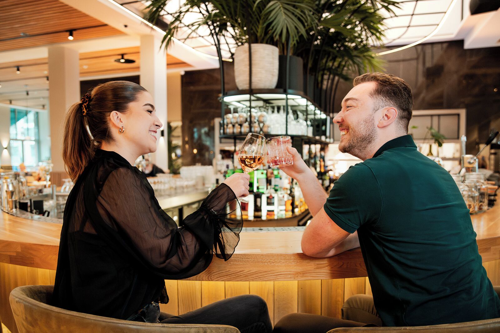 Two people toasting with drinks at a bar with a wooden interior and a large plant in the background.