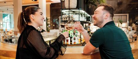 Two people toasting with drinks at a bar with a wooden interior and a large plant in the background.