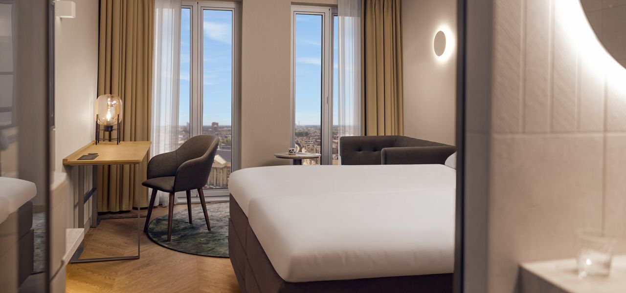 A hotel room with a bed, a desk, a chair, and a view of the city through large windows.