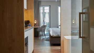 Quarto de hotel moderno com piso de madeira, uma cama, banheiro com banheira, escrivaninha com cafeteira e vista para o exterior.