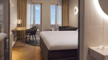 Une chambre d’hôtel avec un grand lit, un bureau avec une chaise et deux fenêtres donnant sur la ville.