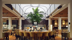 Restaurante moderno com balcão central, pé-direito alto e uma grande planta em interior.