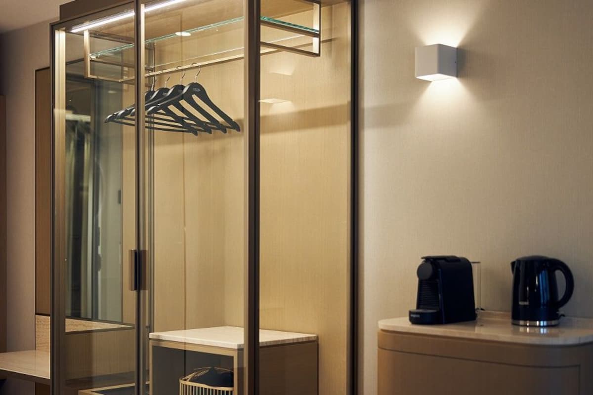 A hotel room with a glass-enclosed wardrobe, a coffee maker on the counter, and a wall-mounted light.