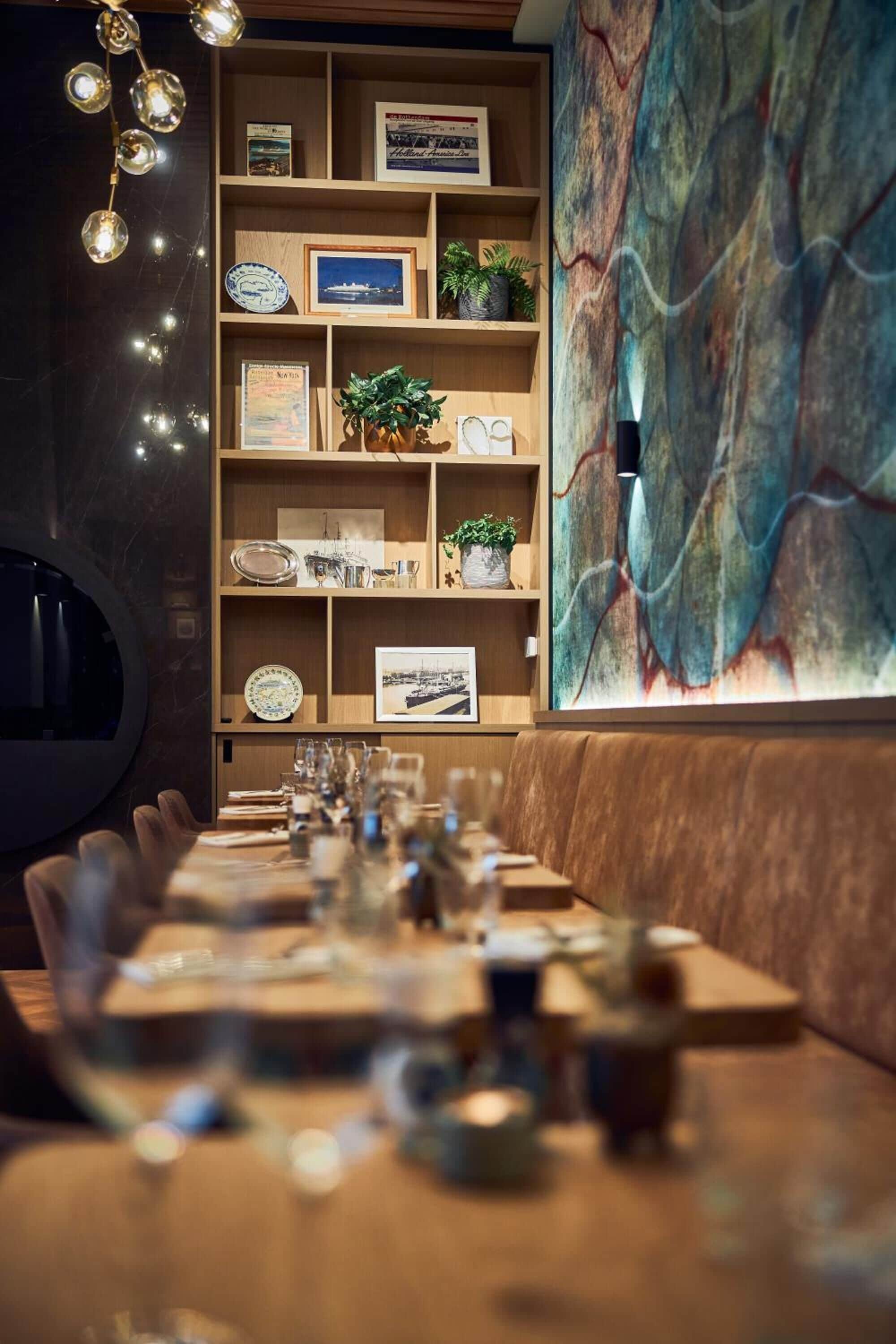 Een restaurant met een lange houten tafel gedekt voor een maaltijd, een houten boekenkast met diverse objecten, en een groot abstract schilderij aan de muur.