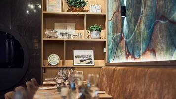 Een restaurant met een lange houten tafel gedekt voor een maaltijd, een houten boekenkast met diverse objecten, en een groot abstract schilderij aan de muur.