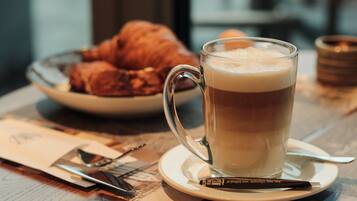 A glass of coffee on a saucer with a croissant in the background.