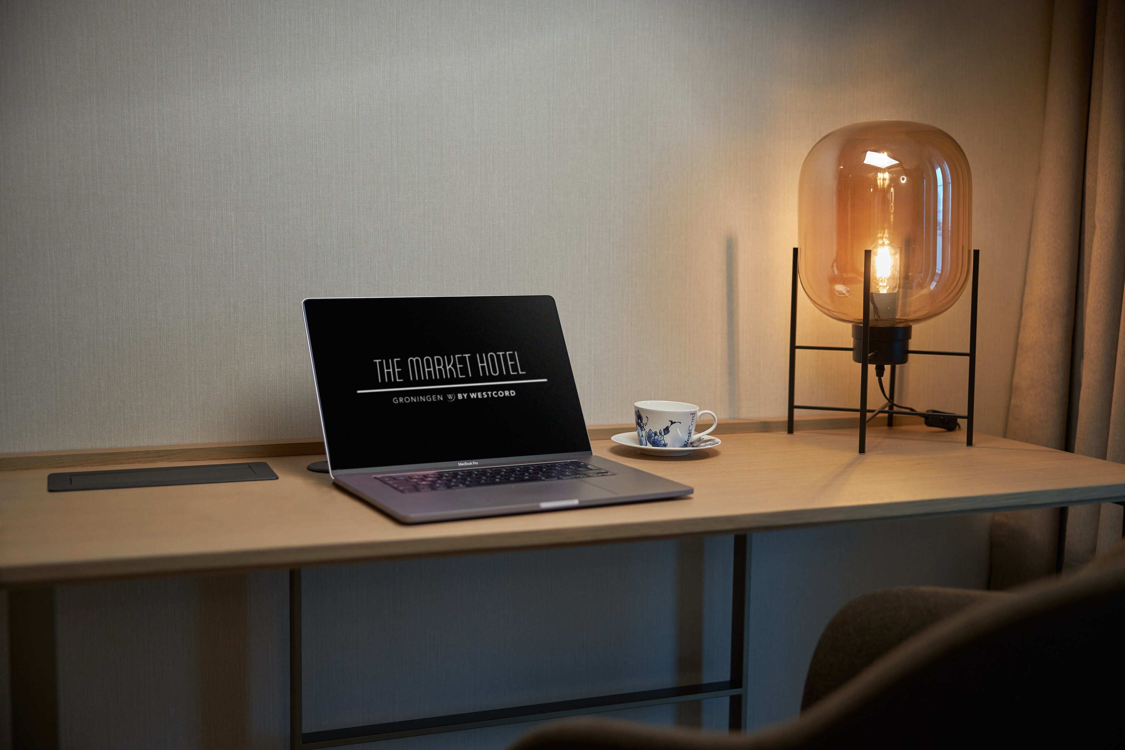Un ordenador portátil sobre un escritorio, una taza al lado, una lámpara con pantalla de cristal transparente y una pared con textura discreta.