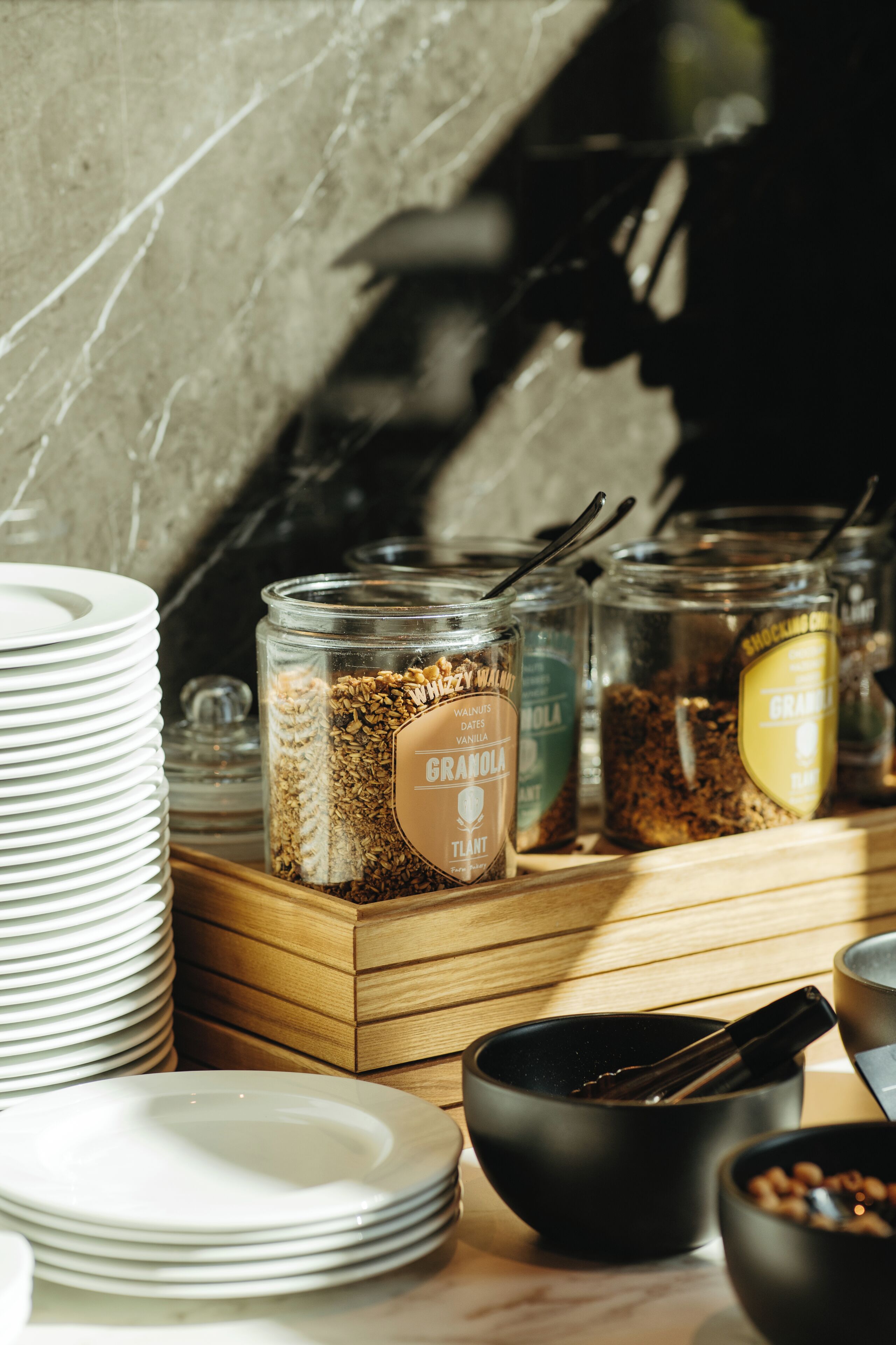 Een ontbijtbuffet met diverse etenswaren in glazen potten, stapels borden ernaast, en een houten serveerplank.