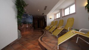 A sunlit indoor area with a tiled floor, a wall-mounted clock, and a row of yellow-striped lounge chairs. - Hotel Grieserhof (Lermoos)