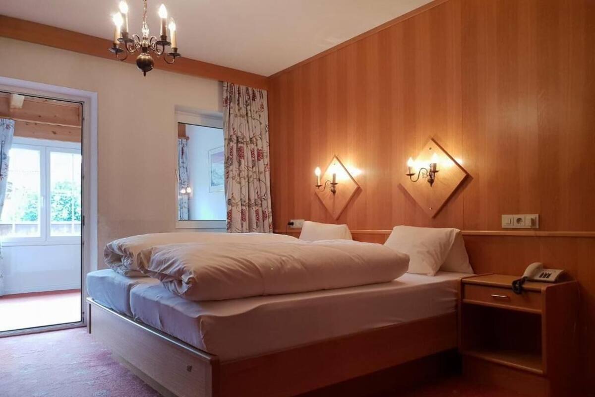 A bedroom with a wooden headboard, a large bed with white linens, a bedside table with a lamp, and a window with curtains.