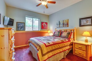 Une chambre à coucher comprenant un lit, une table de chevet en bois, un ventilateur de plafond, un téléviseur fixé au mur et des œuvres d’art accrochées aux murs.