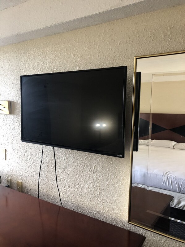 A flat-screen TV mounted on a textured wall, a wooden table in the foreground, and a mirrored wardrobe reflecting a bed with white bedding.