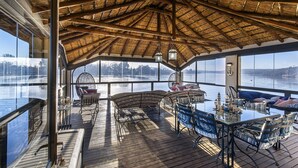 A spacious deck with a thatched roof, large windows offering views of a lake, and a table set for dining. - Vaal River YOLO Spaces – Vaal River Bush Lodge (Metsimaholo)