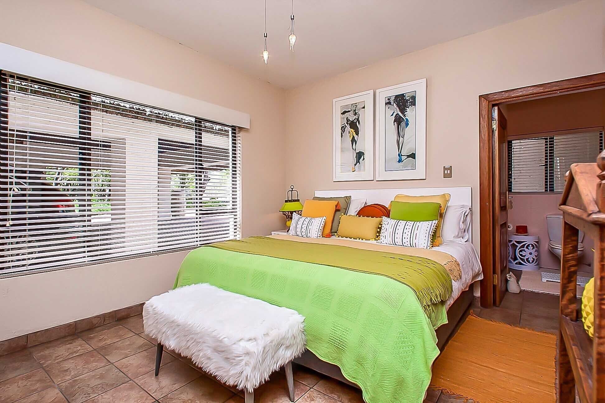 A bedroom with a bed, a bench, a window with blinds, and artwork on the wall.