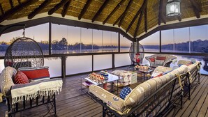A cozy cabin with a thatched roof, large windows, a hanging light fixture, and a cushioned seating area with a view of a lake. - Vaal River YOLO Spaces – Vaal River Bush Lodge (Metsimaholo)