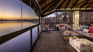 A covered outdoor deck with a view of a lake at sunset, furnished with chairs and a sofa. - Vaal River YOLO Spaces – Vaal River Bush Lodge (Metsimaholo)