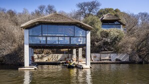 A thatched-roof structure with a glass viewing platform over water, surrounded by trees. - Vaal River YOLO Spaces – Vaal River Bush Lodge (Metsimaholo)