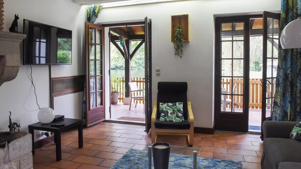 A living room with a TV, a chair, a table, and a view of the outside patio. - Gite Nature et Détente 6/8p with private jacuzzi (Marions)