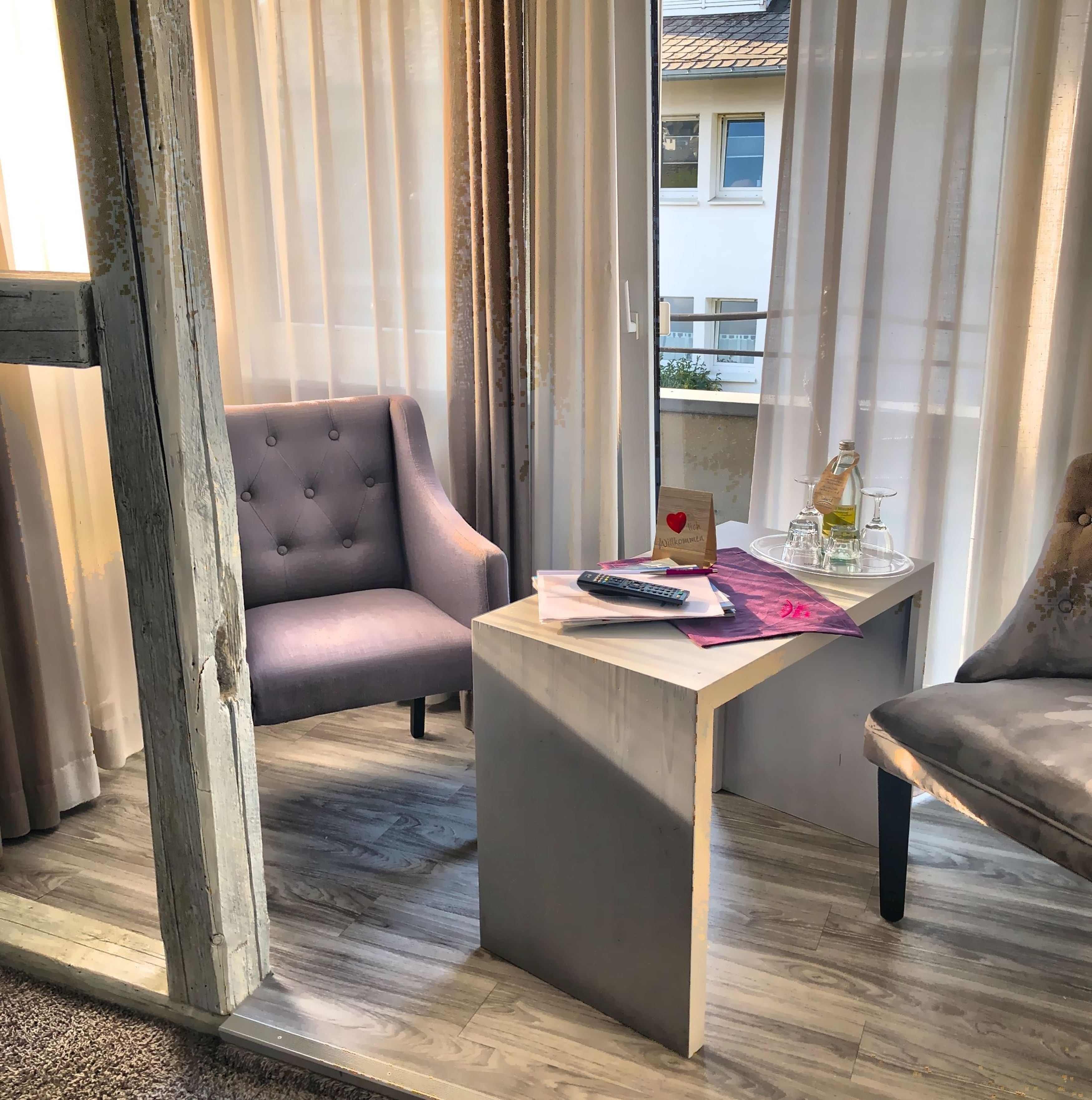 Een kamer met een grijze fauteuil, een klein tafeltje met een afstandsbediening en een uitzicht op gebouwen door dunne gordijnen.