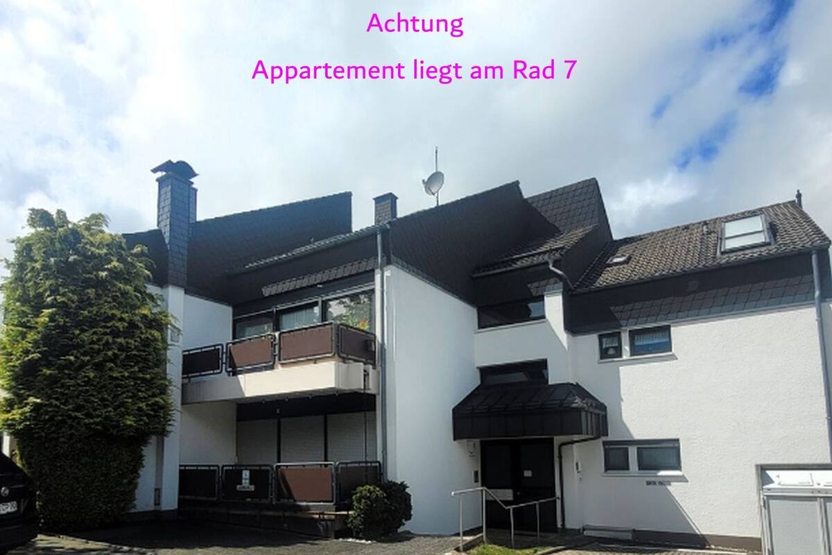 A modern two-story building with a flat roof, a balcony on the upper floor, and a small garden area in front.