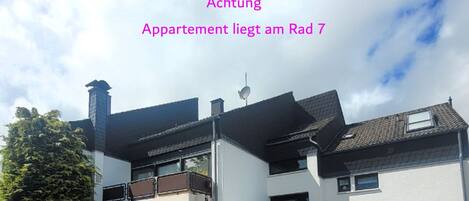 Un bâtiment moderne à deux étages, doté d’un toit plat, d’un balcon à l’étage supérieur et d’un petit jardin à l’avant.