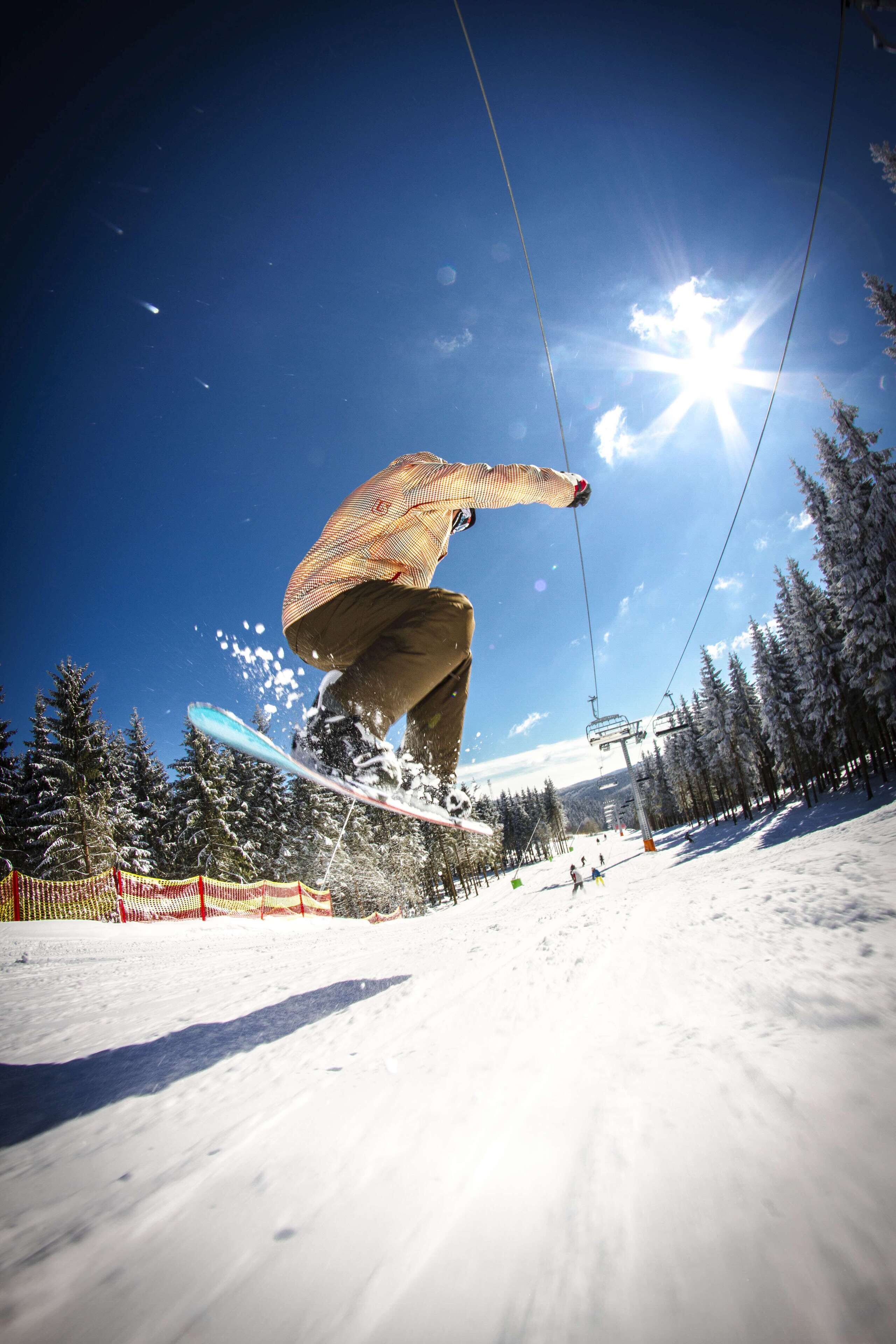 Un snowboarder en pleno vuelo contra un cielo azul despejado, con un efecto de luz solar, rodeado de árboles cubiertos de nieve y un telesilla al fondo.