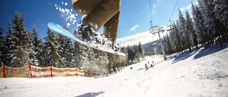 Un snowboarder en pleno vuelo contra un cielo azul despejado, con un efecto de luz solar, rodeado de árboles cubiertos de nieve y un telesilla al fondo.