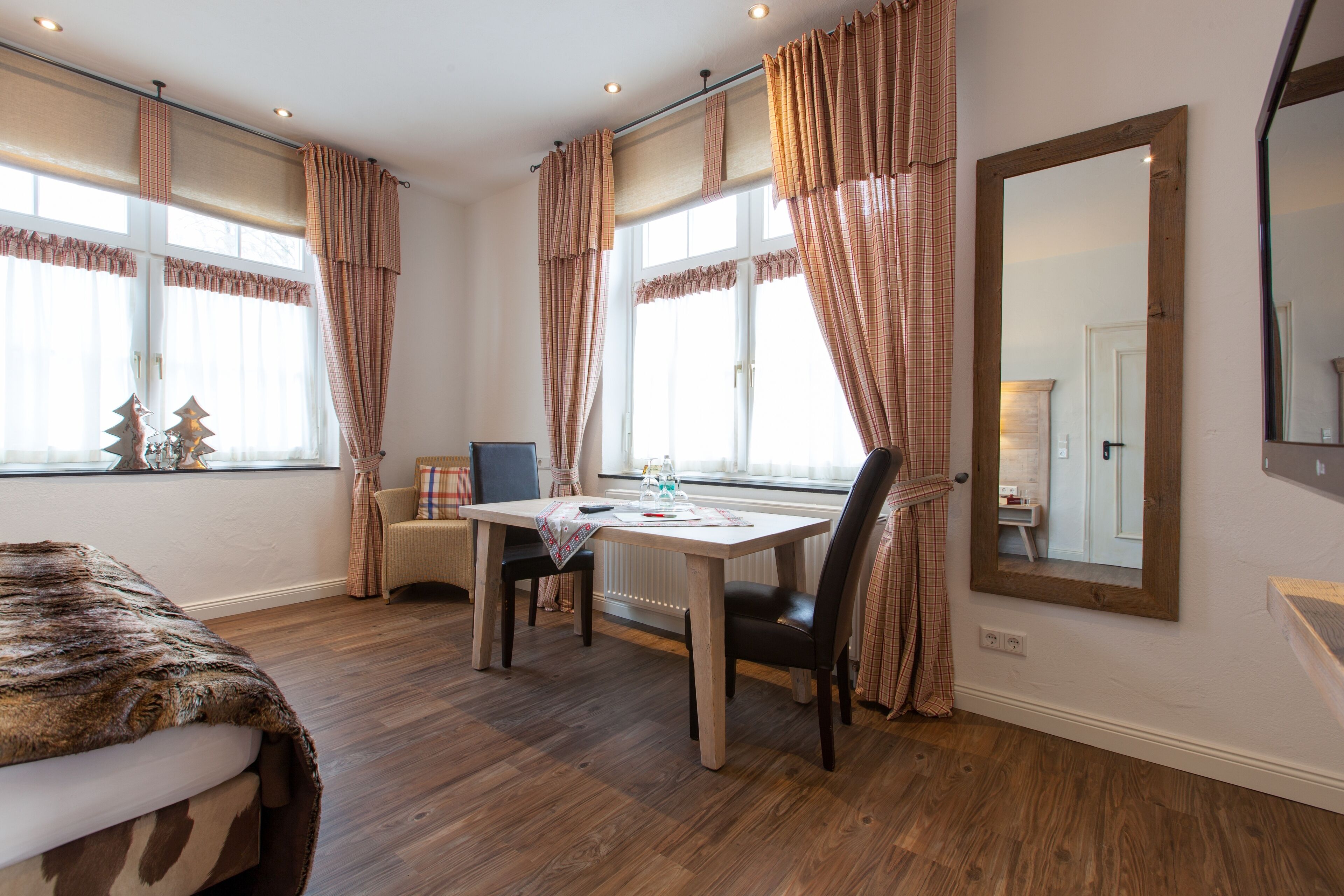 Ein Zimmer mit Holzboden, einem Esstisch mit Stühlen, einem großen Spiegel und Vorhängen.