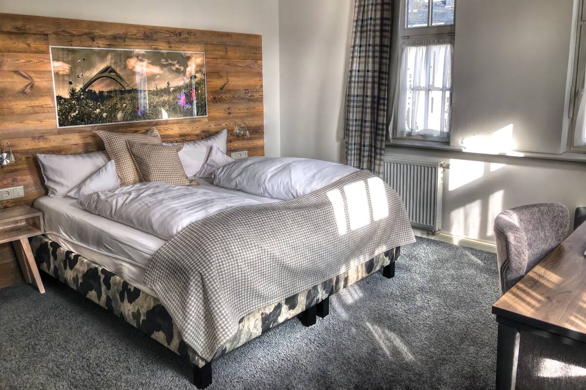A bedroom with a bed, a nightstand, a chair, a window with curtains, and a wall-mounted artwork.