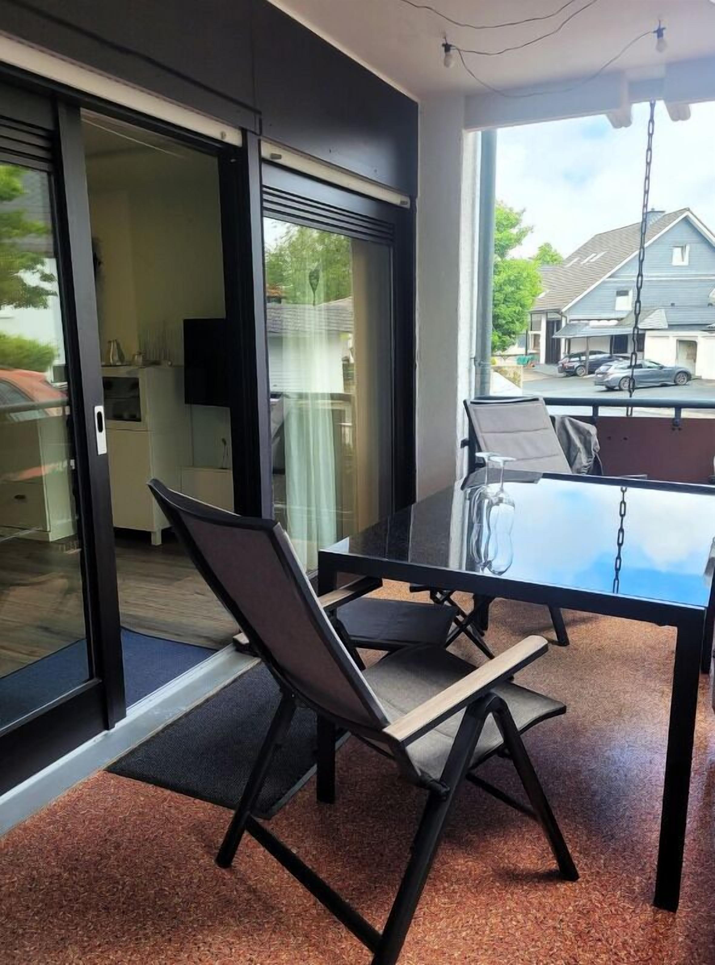 A modern dining area with a glass table and chairs, a sliding glass door leading to a kitchen, and a view of a residential street.