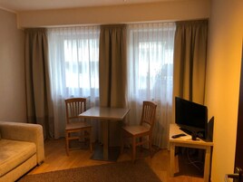Una habitación con sofá, una mesa y dos sillas, televisor sobre un mueble bajo y cortinas de tela ligera.