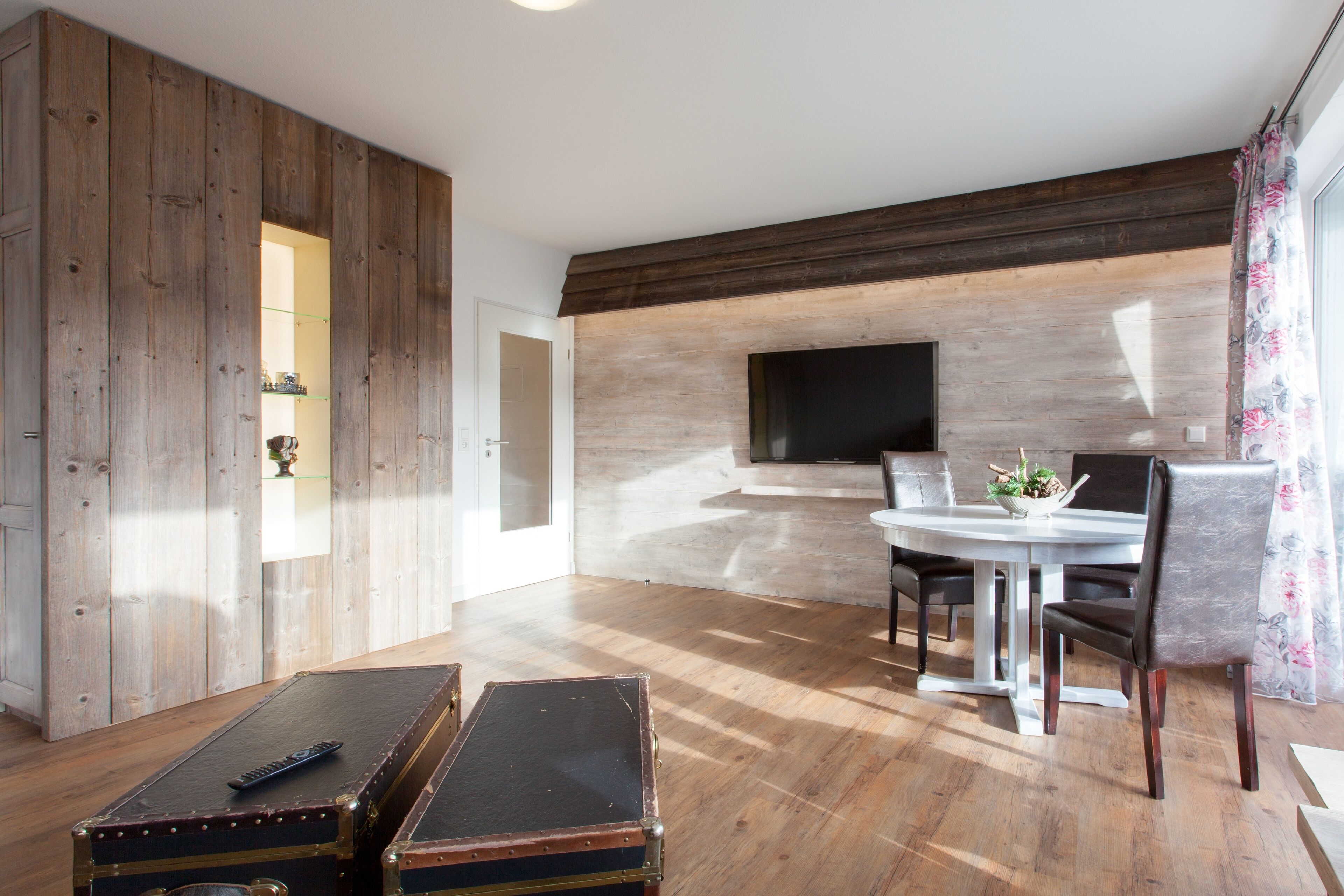 Un salon spacieux doté d’un plancher en bois, d’une table à manger avec des chaises, d’un téléviseur fixé au mur et d’une grande armoire en bois.