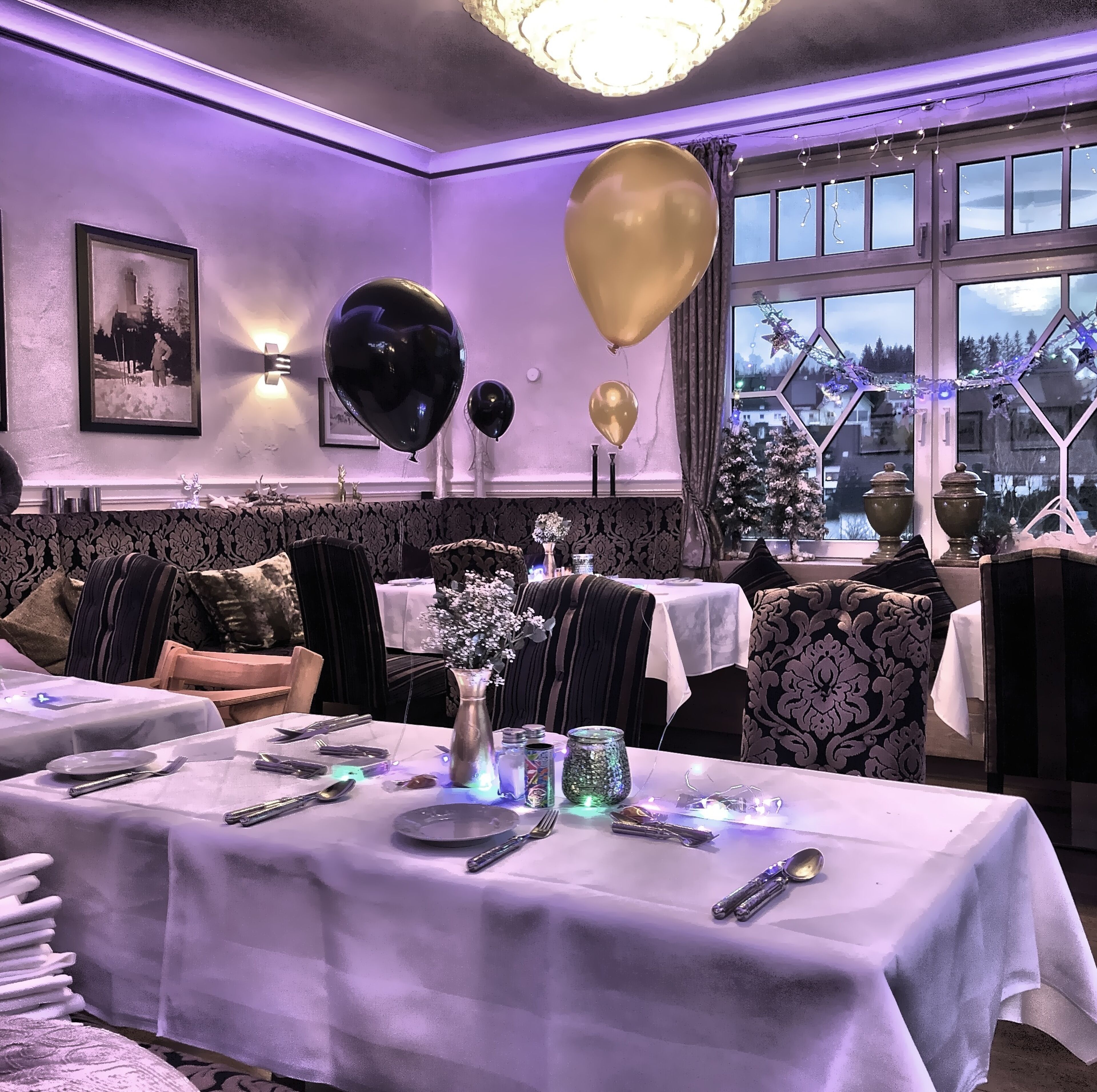 Een eetruimte met gedekte tafels, met een kroonluchter, zwarte en gouden ballonnen en een uitzicht op het buitenleven door grote ramen.