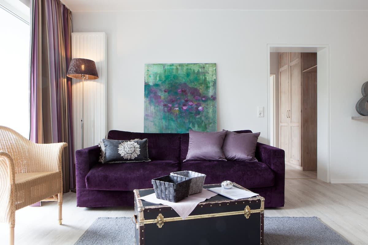 A living room with a purple sofa, a wicker chair, a black suitcase, and a painting on the wall.